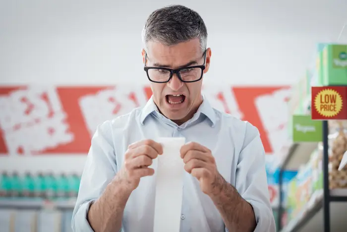 This is a stock photo of a man in glasses, looking shocked while examining a long receipt.
